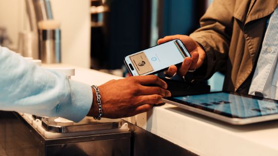 Eine Person hält an einem Tresen ein Smartphone, auf dessen Bildschirm eine American Express Kreditkarte abgebildet ist, an ein Kartenlesegerät, das ihr eine andere Person hinhält. 
