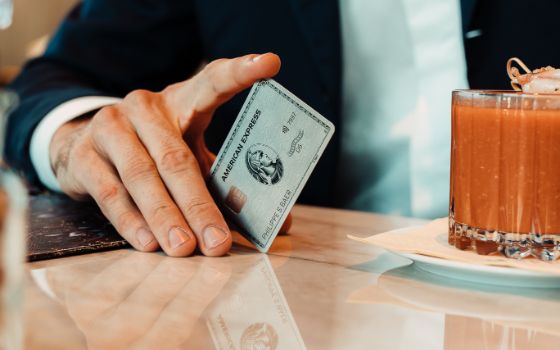 Nahaufnahme einer unkenntlichen Person im Anzug, die die Hand auf einen Tisch gestützt hat und mit einem Finger eine silberne Kreditkarte balanciert, daneben ein Cocktailglas auf einem weissen Teller.