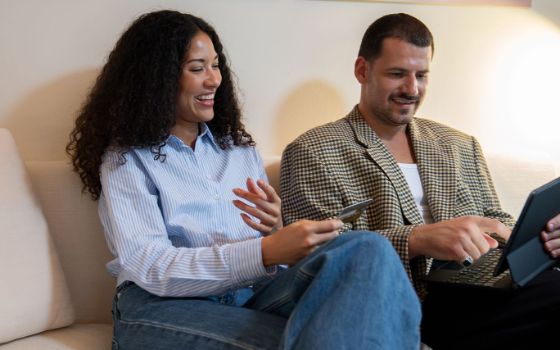Eine Frau und ein Mann sitzen gut gelaunt auf einem Sofa, gemeinsam schauen sie auf ein Tablet, das er auf seinem Schoss bedient, sie hält eine Kreditkarte in der Hand. 