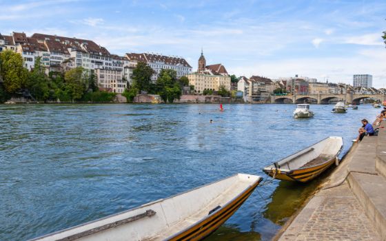Blick auf die Basler Altstadt mit Mittlerer Brücke, Rheinpromenade und traditionellen Fähri-Booten bei sommerlichem Wetter.