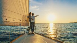 Rückansicht eines sich umarmenden Paares am Bug einer Segeljacht auf dem Meer bei Sonnenuntergang.