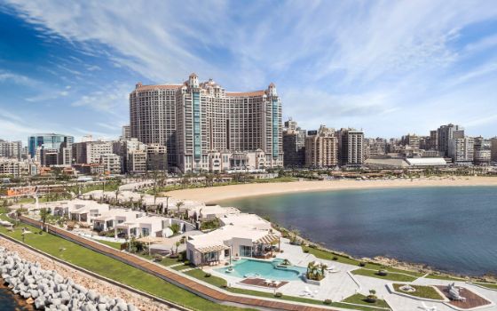 Hotelkomplex an einem Sandstrand bei Sonnenschein, im Vordergrund Cabanas mit Poollandschaft auf einer Landzunge. 