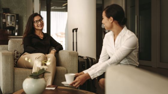 Eine Frau sitzt in einem Sessel und lächelt eine Kellnerin an, die ihr Kaffee serviert, neben ihr steht ein Rollkoffer.