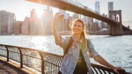 Eine Frau in Jeansjacke steht vor einer Brücke an einem Geländer am Flussufer vor der Skyline von Manhattan und macht lächelnd ein Selfie mit einem Smartphone bei Sonnenschein. 