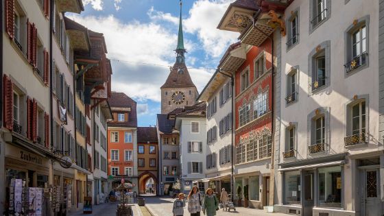 Personen laufen durch eine Fußgängerzone, umgeben von mittelalterlichen Gebäuden mit bemalten Giebeln und Kirchturm in der Altstadt von Aarau. 