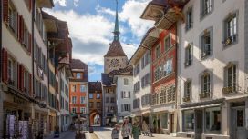 Personen laufen durch eine Fußgängerzone, umgeben von mittelalterlichen Gebäuden mit bemalten Giebeln und Kirchturm in der Altstadt von Aarau. 