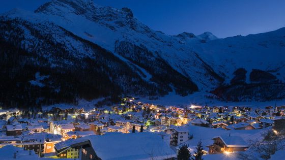 Vor einer dunklen Berglandschaft bei Nacht befindet sich ein kleines Dorf mit etlichen beleuchteten Häusern. 