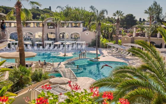 Personen in der Poollandschaft einer Hotelanlage mit Palmen bei Sonnenschein. 