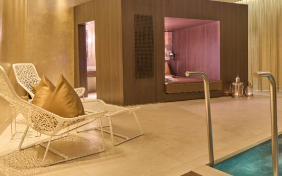 Ein indirekt beleuchteter Raum mit Steinboden, zwei weissen Liegestühlen, einem Whirlpool und einer Sauna mit Holzverkleidung. 