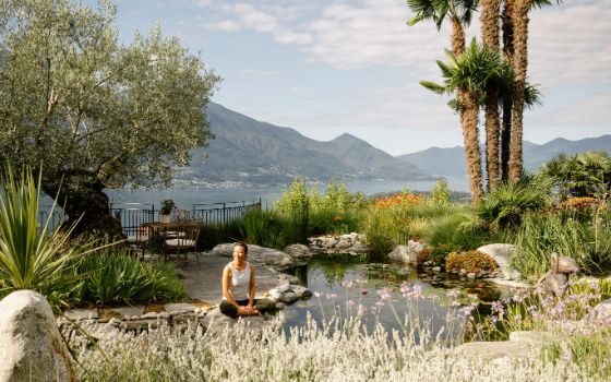 Eine Frau sitzt lächelnd in einem mediterranen Garten an einem Teich, im Hintergrund ein See und Bergpanorama. 