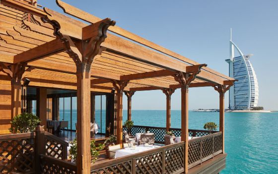 Eine Terrasse aus Holz mit gedeckten Tischen ragt über das blaue Wasser, im Hintergrund sieht man das Gebäude Burj Al Arab. 