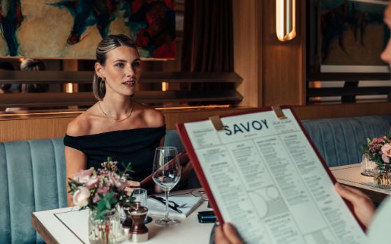 Eine elegante Frau im schwarzen Abendkleid sitzt an einem Tisch in einem edlen Restaurant einem Mann mit grosser Speisekarte in den Händen gegenüber.