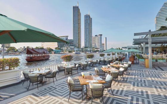 An einem Fluss stehen auf einer Terrasse gedeckte Tische, im Hintergrund ragen Hochhäuser empor.