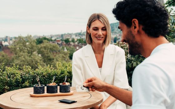 Ein lächelndes Paar sitzt an einem Tisch auf einer Dachterrasse, er hält eine silberne Kreditkarte von American Express in der Hand. 