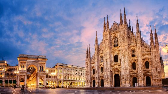 Der Mailänder Dom mit zahlreichen verzierten Türmchen und Fenstern steht direkt neben dem Torbogen des Eingangs einer beleuchteten Galerie, hinter den Gebäuden geht die Sonne unter. 
