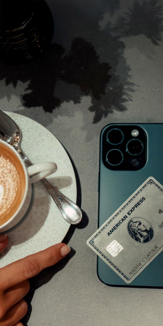 Eine Hand greift nach einer Tasse Cappuccino, die auf einer grauen Tischplatte steht, daneben liegt ein Smartphone mit einer Kreditkarte von American Express.