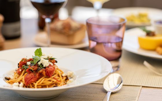 Spaghetti mit Tomaten und Basilikum sind auf einem weissen Teller angerichtet, der steht auf einem Holztisch mit farbigen Gläsern davor. 