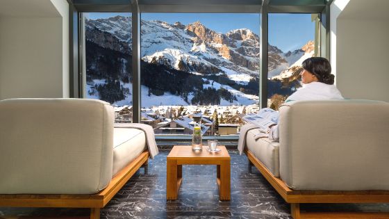 Rückansicht einer Frau im weissen Bademantel auf einer gepolsterten Liege vor einem Panoramafenster mit Blick auf eine verschneite Berglandschaft.