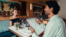 Ein elegant gekleidetes Paar sitzt sich gegenüber an einem Tisch in einem stilvollen Restaurant und hält Speisekarten in den Händen.