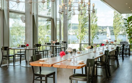 Heller Gastraum eines Restaurants mit langer Holztafel, an der auf beiden Seiten viele schwarze Stühle stehen, vor einer grossen Fensterfront, im Hintergrund liegt der Zürichsee, auf dem ein Segelboot treibt.