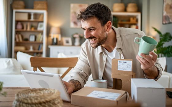Ein Mann hält eine Tasse in der Hand, vor ihm liegen auf einem Tisch Pakete und ein Laptop. 