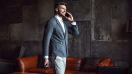 Mann im Anzug telefoniert lächelnd und zieht einen Rollkoffer hinter sich her, im Hintergrund ein Ledersofa.