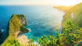 Am Kelingking Strand auf Bali ragt ein grosser Berg neben eine Bucht mit blauem Meer und Sandstrand auf. 