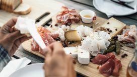 Zwei Hände mit Messer an einem angerichteten Holzbrett mit Wurst und Käse auf einem Holztisch.