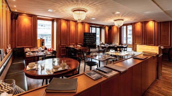 Eleganter Speisesaal mit Holzverkleidung und bodentiefen Fenstern, Deckenlampen aus Kristall und eingedeckten Holztischen.