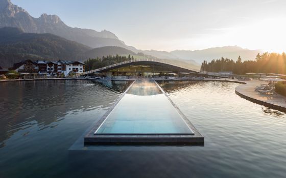 Modernes Hotelresort mit geschwungenem Dach und einem langgezogenen Infinity-Pool in einem Teich, im Hintergrund Bergpanorama bei tiefstehender Sonne. 