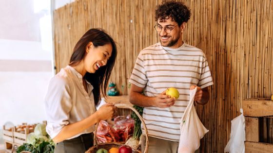 Ein junges Paar steht vor einem Marktstand, sie hält einen Einkaufskorb mit Obst und er eine Tragetasche. 