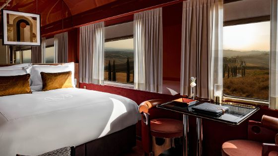 Innenaufnahme eines luxuriösen Zugwaggons mit roten Wänden, eingerichtet mit Tisch und Doppelbett. Blick durch mehrere Fenster mit geöffneten Gardinen auf eine sanfte Hügellandschaft mit Zypressen.