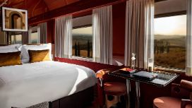 Innenaufnahme eines luxuriösen Zugwaggons mit roten Wänden, eingerichtet mit Tisch und Doppelbett. Blick durch mehrere Fenster mit geöffneten Gardinen auf eine sanfte Hügellandschaft mit Zypressen.