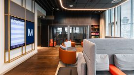 Eine Flughafen-Lounge am Airport München mit Holzboden, Sofas, Sesseln, einem Flugmonitor und Fensterfront zur Rollbahn.