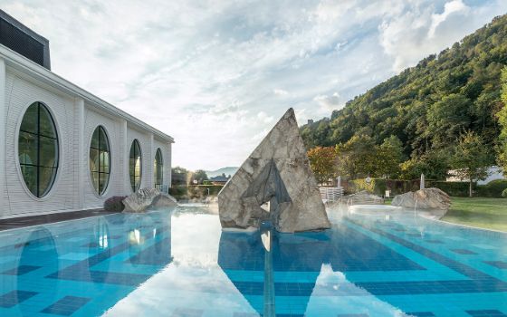 In einem Aussenpool steht mittig im Wasser eine Marmorskultptur, links daneben steht ein weisses Gebäude, umgeben ist der Pool von einer grünen Landschaft. 