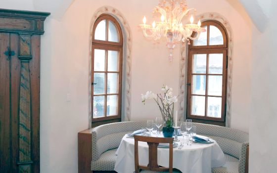 In einem kleinen Erker mit weissen Wänden und schmalen rundzulaufenden Fenstern ist eine runde Sitzecke, an dem ein Tisch gedeckt ist. An der Decke hängt ein Kronleuchter. 