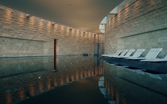 In einem Spa mit beigen Steinwänden ist ein großer Indoor-Pool, an dem weisse Liegen stehen. 