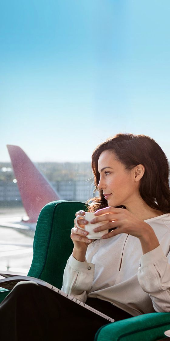 Eine stilvoll gekleidete Frau sitzt mit einer Tasse Kaffee in den Händen in einem grünen Sessel an einer Glasfassade in einer Flughafen Lounge und blickt entspannt hinaus aufs Rollfeld unter blauem Himmel.