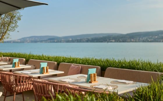 Sofas und Rattanstühle an mehreren Tischen auf einer begrünten Sonnenterrasse bei sonnigem Wetter mit See und Hügeln im Hintergrund.