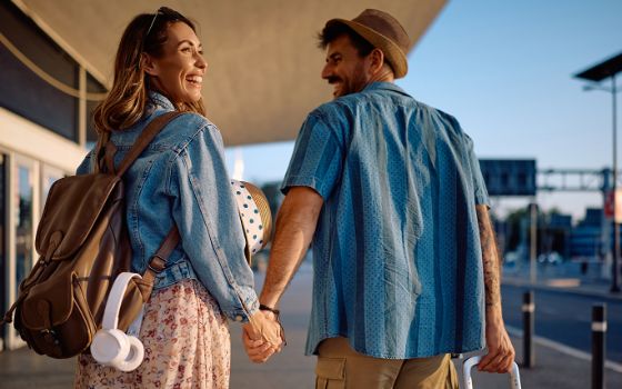 Eine Frau und ein Mann stehen Hand in Hand vor einem Flughafen. 