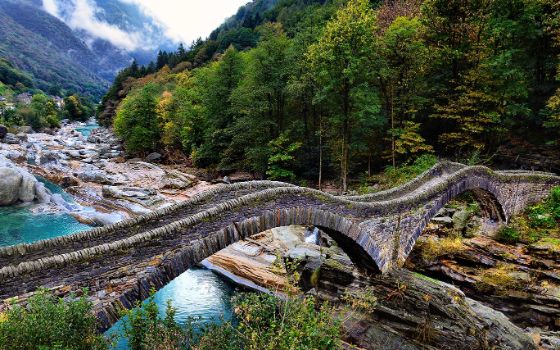 Eine Steinbrücke in Wellenform führt vom Ufer in einen Wald, im Hintergrund sieht man Berge.