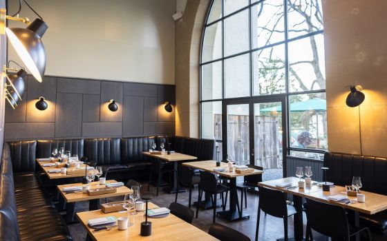 Stilvoller, heller Speisesaal im Brasserie-Stil mit schwarzer Bestuhlung an hellen Holztischen mit Blick auf eine Terrasse durch ein Panoramafenster. 