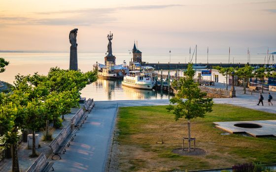 Hafen von Konstanz am Bodensee bei Sonnenuntergang mit der Imperia-Statue, Anlegestellen für Ausflugsschiffe und Baumallee im Vordergrund.
