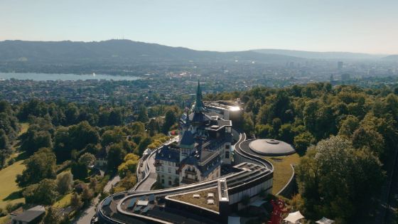 Blick aus der Vogelperspektive auf ein grosses Anwesen mit schlossartigen Türmen inmitten von Bäumen, dahinter eine Stadt und ein See.