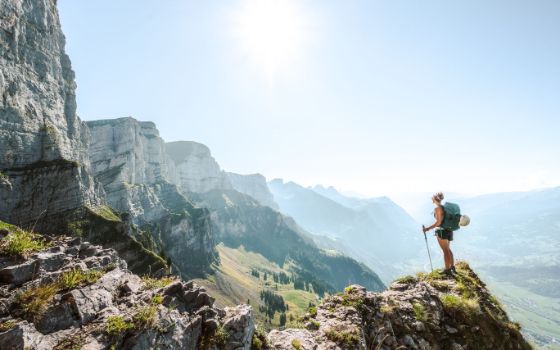 Wanderin mit Rucksack auf Felsvorsprung in den Schweizer Alpen mit Blick ins sonnige Tal bei klarem Himmel