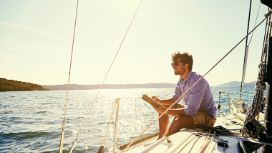 Ein Mann sitzt auf einem Segelboot und fährt auf dem Meer, er hat ein Buch und einen Stift in der Hand und schaut auf die offene See. 