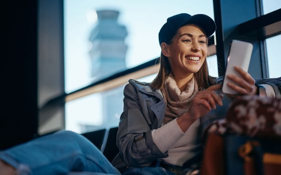 Eine junge Frau sitzt in einer Flughafen-Lounge und schaut auf ihr Smartphone. 
