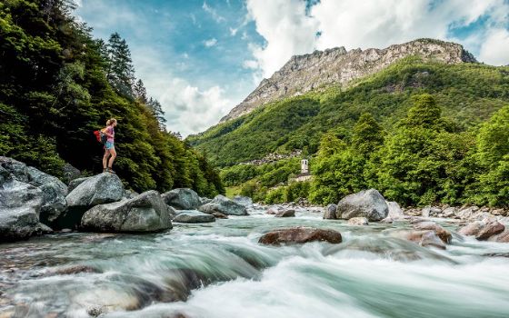 Eine Frau steht auf einem grossen Stein am Fluss von Verzasca, im Hintergrund das Dorf Lavertezzo. 