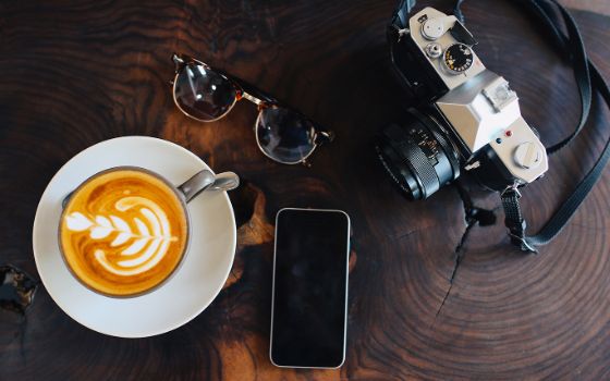 Eine Tasse Cappuccino, eine Sonnenbrille, ein Smartphone und eine Kamera liegen auf einer Tischplatte. 