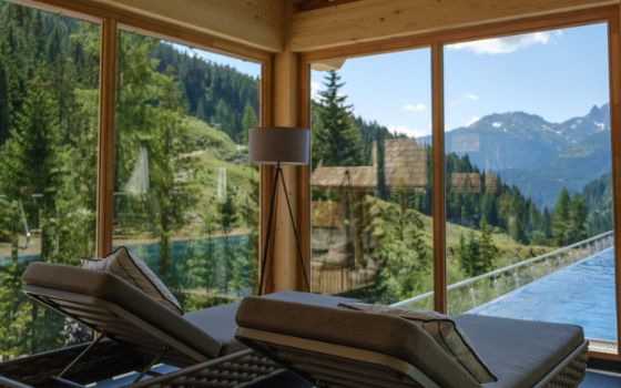 Rückansicht zweier Polsterliegen in einem Raum mit bodentiefen Panoramafenstern mit Blick auf eine bewaldete Berglandschaft und einen Infinity-Pool. 